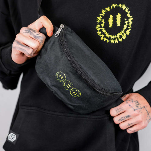 Mann mit Tattoos trägt Technostoff OFF TRACK Hip Bag für Raver mit Acid Smileys in gelb