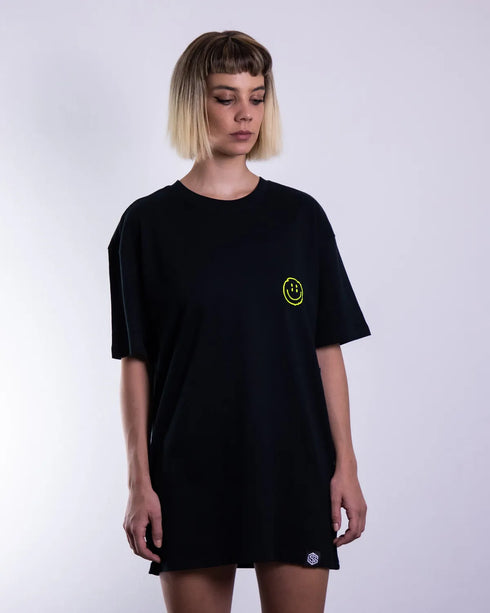 Schwarzes "Distorted Smile" oversized T-Shirt mit neon gelben Smily-Flockdruck auf dem Rücken, getragen von einer Frau, fotografiert von vorne