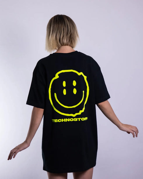 Schwarzes "Distorted Smile" oversized T-Shirt mit neon gelben Smily-Flockdruck auf dem Rücken, getragen von einer Frau, fotografiert von hinten