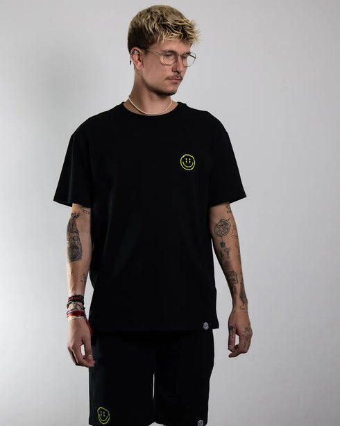 Schwarzes "Distorted Smile" oversized T-Shirt mit neon gelben Smily-Flockdruck auf dem Rücken, getragen von einem Mann, fotografiert von vorne