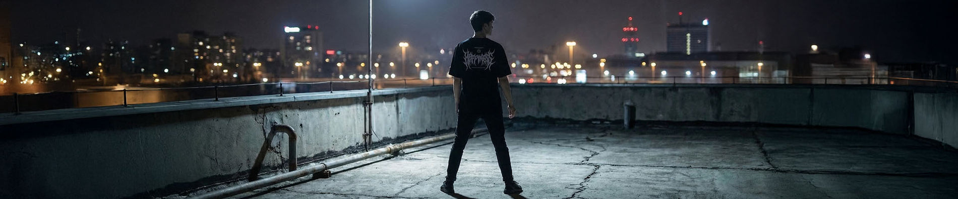 Mann in schwarzem Technostoff Happiness T-Shirt steht auf einem Betonrooftop bei Nacht, Stadtlichter im Hintergrund