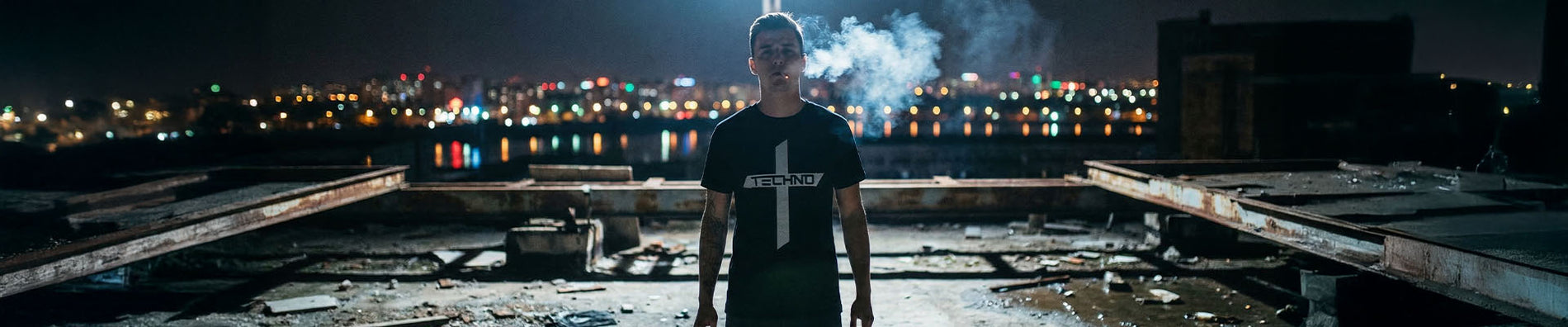 Mann in schwarzem Technostoff Techno Cross T-Shirt steht rauchend auf einem verlassenen Betonrooftop bei Nacht, Stadtlichter im Hintergrund