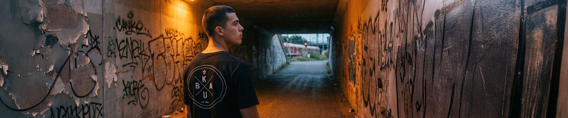 Mann in schwarzem Kuba Warburg Welda T-Shirt steht in einer Graffiti-Unterführung bei Nacht, warmes Licht im Hintergrund