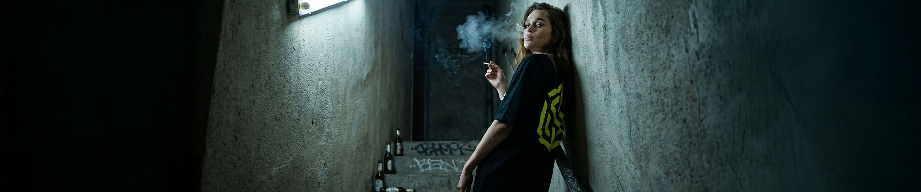 Frau in schwarzem Technostoff Yellow Sign T-Shirt lehnt rauchend an einer Betonwand in einem dunklen Treppenhaus, Graffiti im Hintergrund