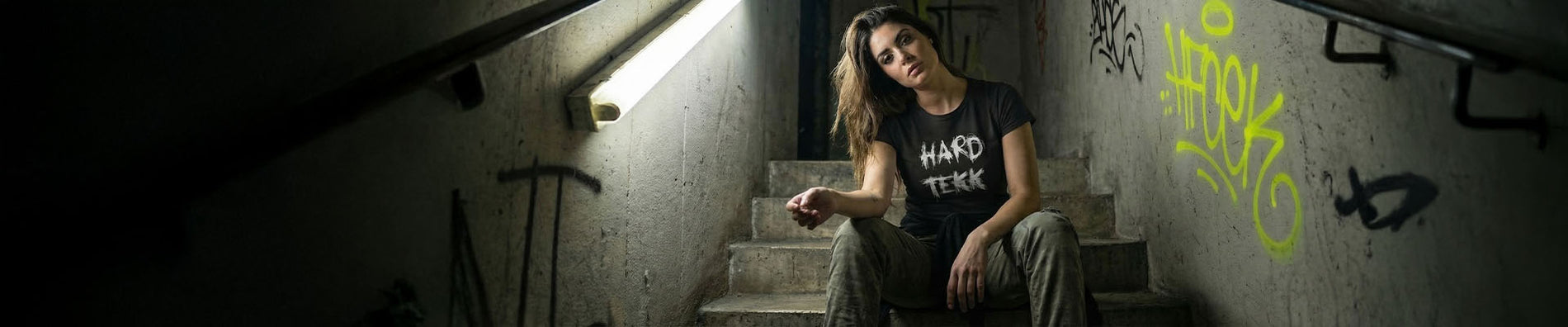 Frau in schwarzem Hard Tekk T-Shirt sitzt auf einer Betontreppe in einem verlassenen Treppenhaus, Neonlicht von links, Graffiti an der Wand