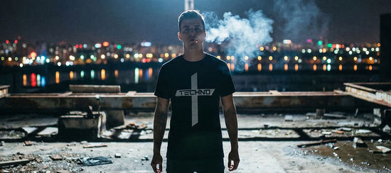 Mann in schwarzem Technostoff Techno Cross T-Shirt steht rauchend auf einem verlassenen Betonrooftop bei Nacht, Stadtlichter im Hintergrund