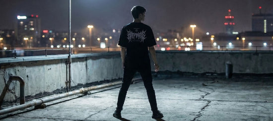 Mann in schwarzem Technostoff Happiness T-Shirt steht auf einem Betonrooftop bei Nacht, Stadtlichter im Hintergrund