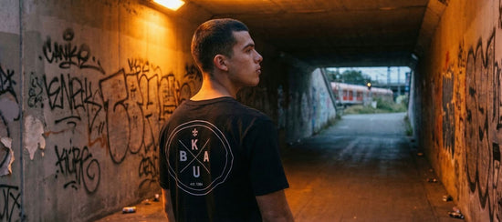 Mann in schwarzem Kuba Warburg Welda T-Shirt steht in einer Graffiti-Unterführung bei Nacht, warmes Licht im Hintergrund