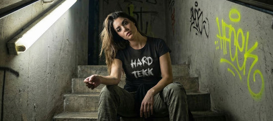 Frau in schwarzem Hard Tekk T-Shirt sitzt auf einer Betontreppe in einem verlassenen Treppenhaus, Neonlicht von links, Graffiti an der Wand