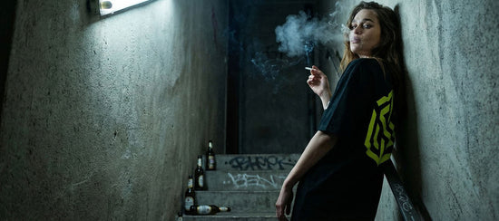 Frau in schwarzem Technostoff Yellow Sign T-Shirt lehnt rauchend an einer Betonwand in einem dunklen Treppenhaus, Graffiti im Hintergrund